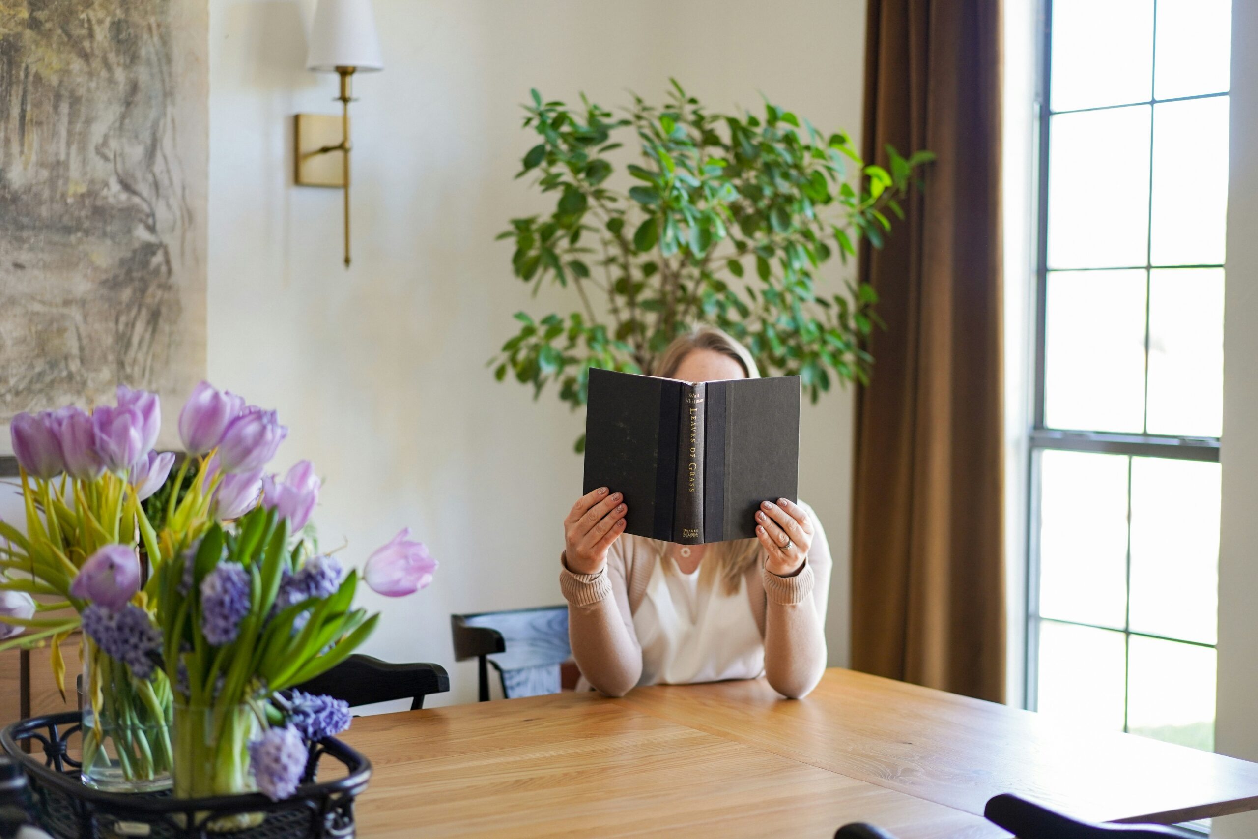 Person reading at table, book is covering their face, there are floral arrangements around them