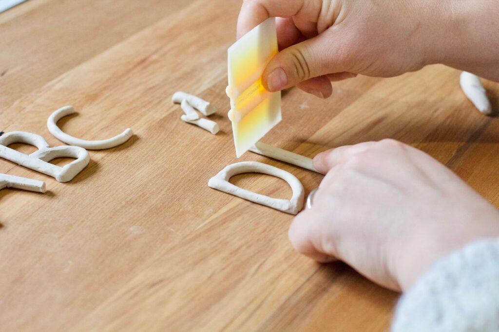Clay letter D on wooden board with hands cutting another letter next to the D