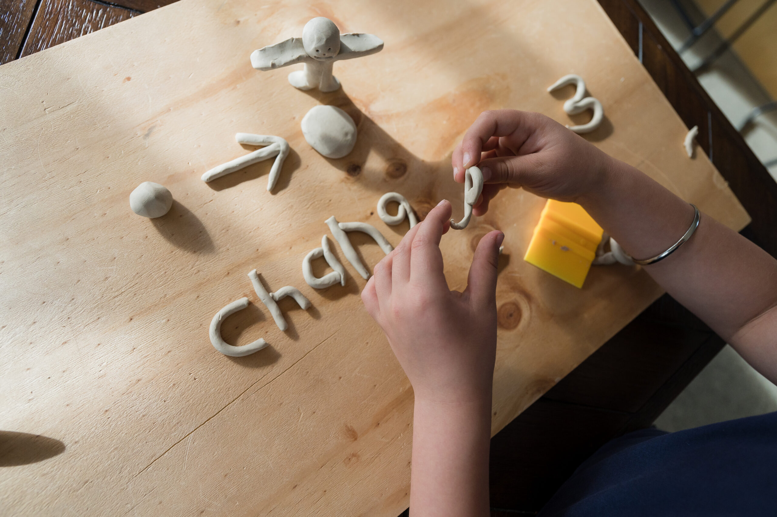 Hands creating the letter "e" with clay over a wooden table. On the table the word change has been spelled out in clay minus the "e". Above change is a clay ball, with an arrow pointing to a flattened ball, with a clay person stnading next to it.