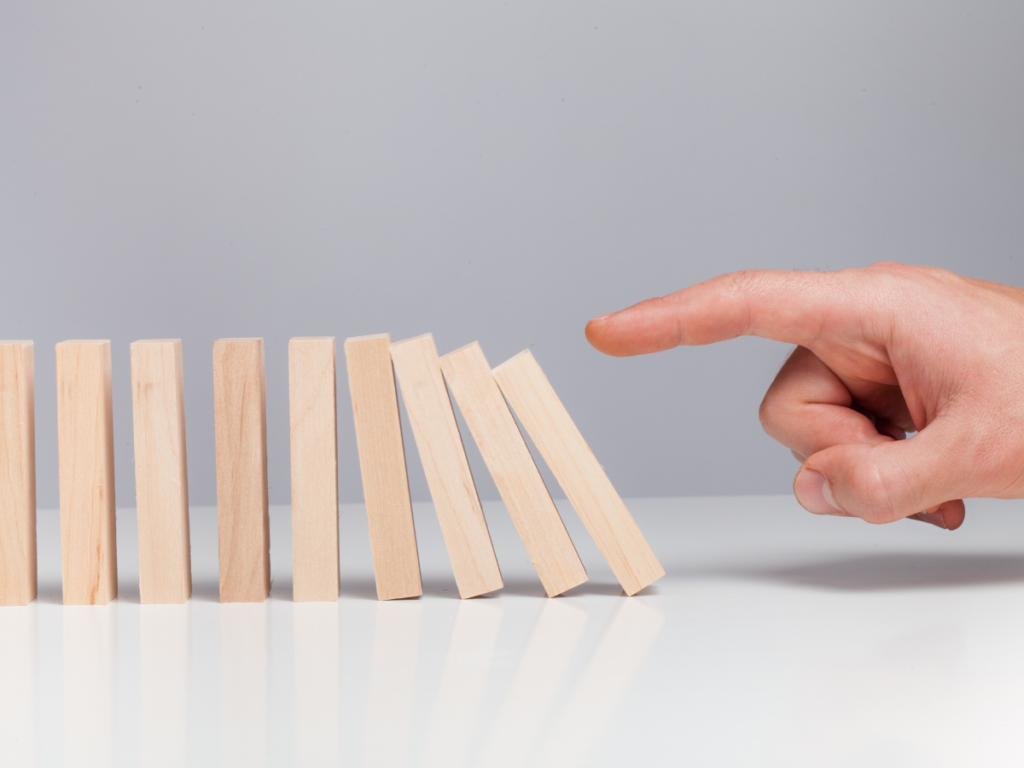 Dominos lined up with a finger pointing at them, the finger has just pushed the dominos, the first three are touching each other with the fourth falling onto the fifth. There are 4 more dominos in the line that will soon fall