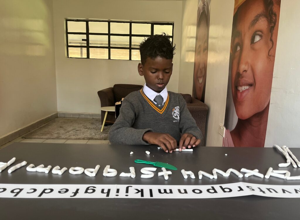 A young boy in a school uniform with the alphabet modelled out of clay and on paper in front of him