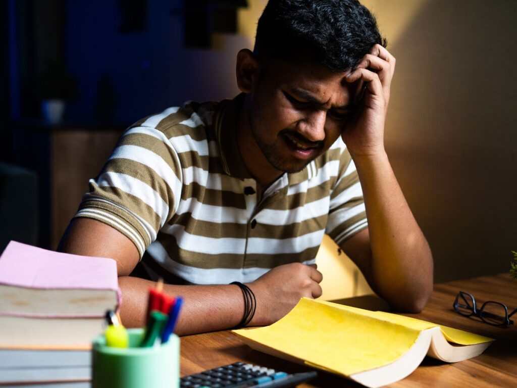 Image of a man in his late 20s, looking pained, with a book open, but face down on a table. He looks to be upset he is having difficulty reading