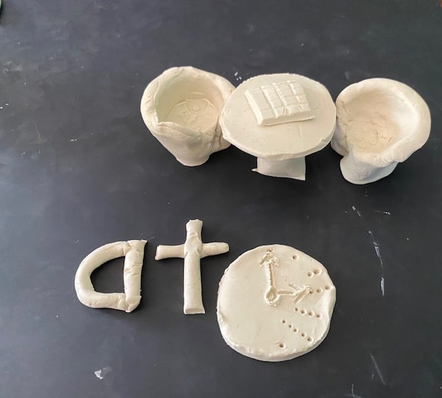 A table with chocolate on it, 2 chairs the word "at" and a clock showing 3 o'clock, all modelled from clay as part of the Davis Mastery for Dyslexia program