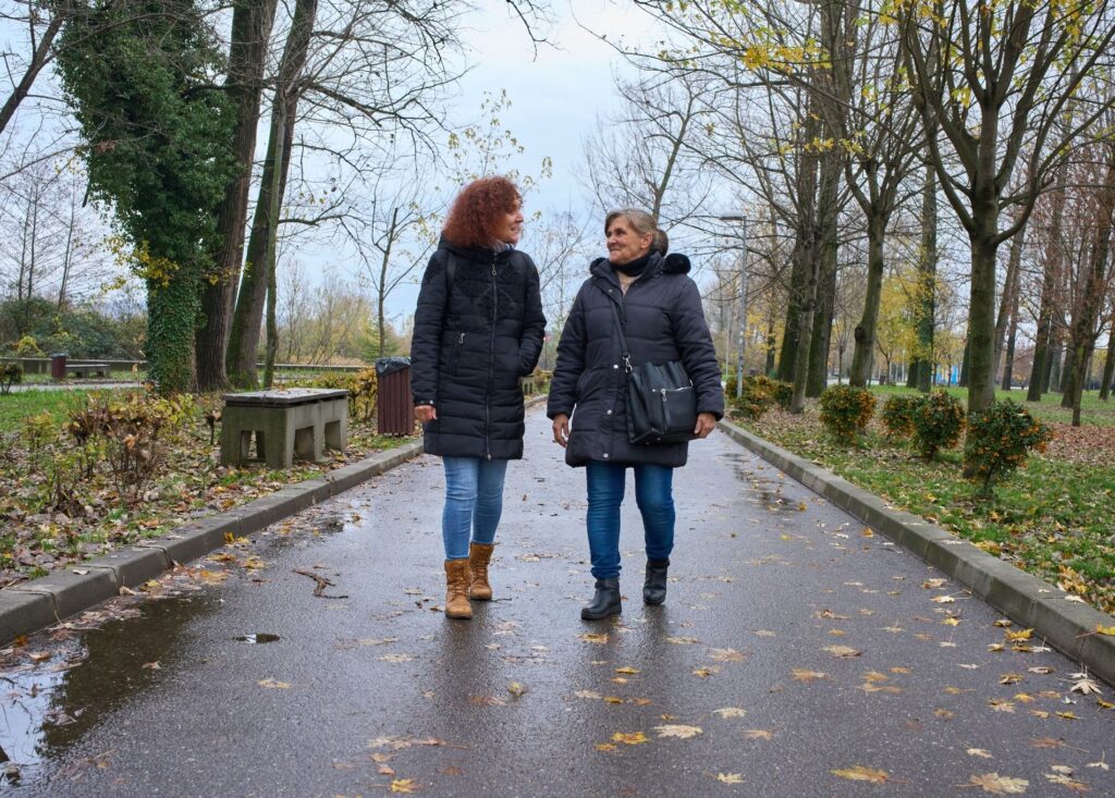 wo women, walking outside. the weather looks like it has been raining but is now clearing. The path is wet and there are fallen leaves around them