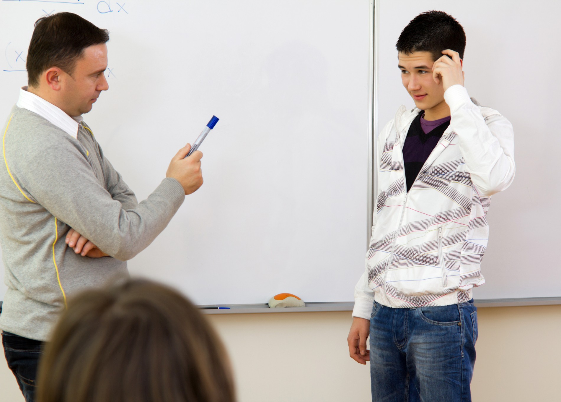 A teacher and student stnading in front of a whiteboard, teacher is holding out the pen - student looks overwhelmed and upset