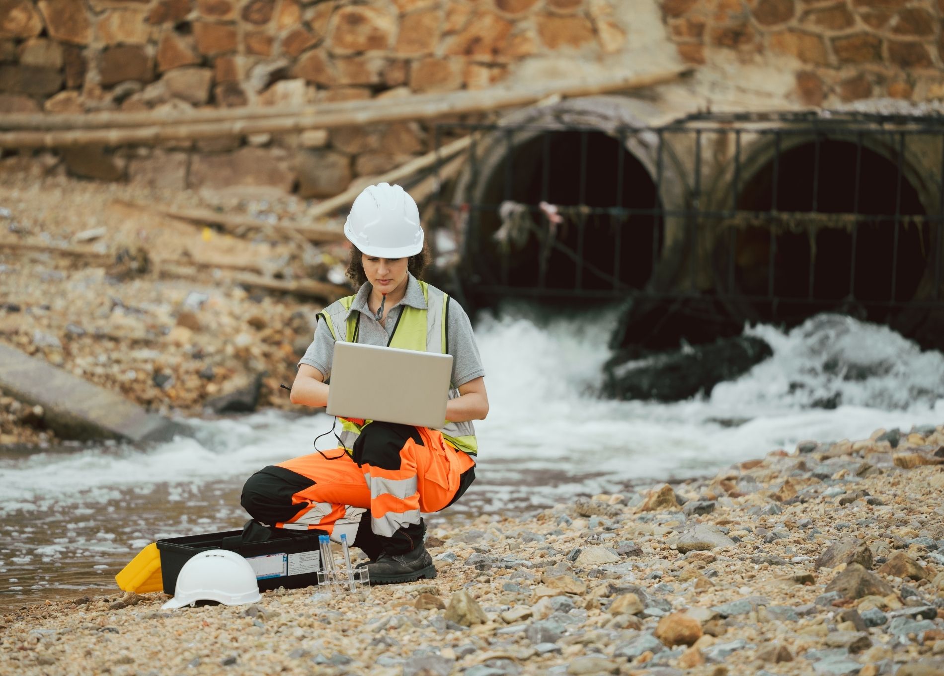 Environmental scientist in the field. Sitting in front of a brick over bridge with two arches. There water running through them. The female scientist is wearing high vis and has her hard ahad and a lab kit besider her. She is crouching with a laptop on her knee